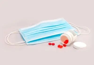 A surgical mask next to an open pill bottle that&rsquo;s toppled over, spilling out red capsules meant to represent vitamin D supplements.